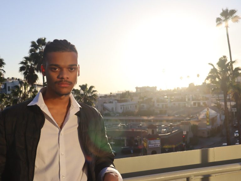 Caleb Morris posed on top of a rooftop overlook facing the camera. Dressed in a long sleeve white collared shirt under a black puffer jacket. The building overlooks a sunset on Hollywood Boulevard